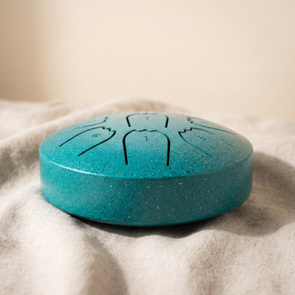 Teal steel tongue rain drum on natural linen, showing the nine cut tongues