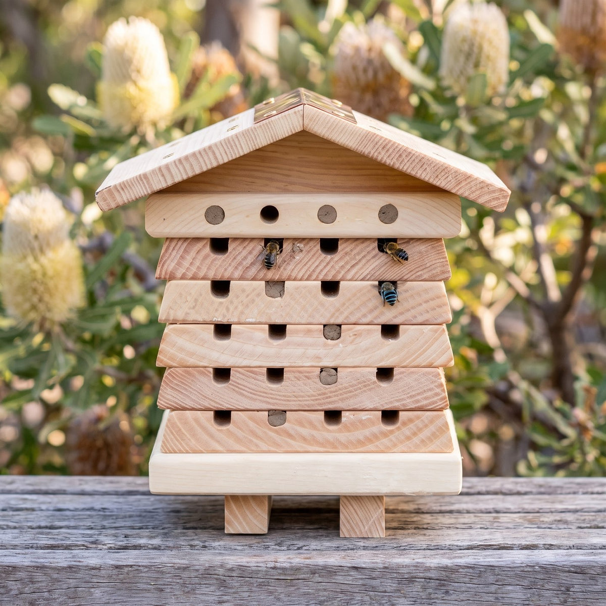 Hollow Keeper bee hotel on a timber garden bench: five stacked cedar boards with notched nesting openings under a shallow cedar gable roof with a narrow copper ridge strip, native bees at the openings